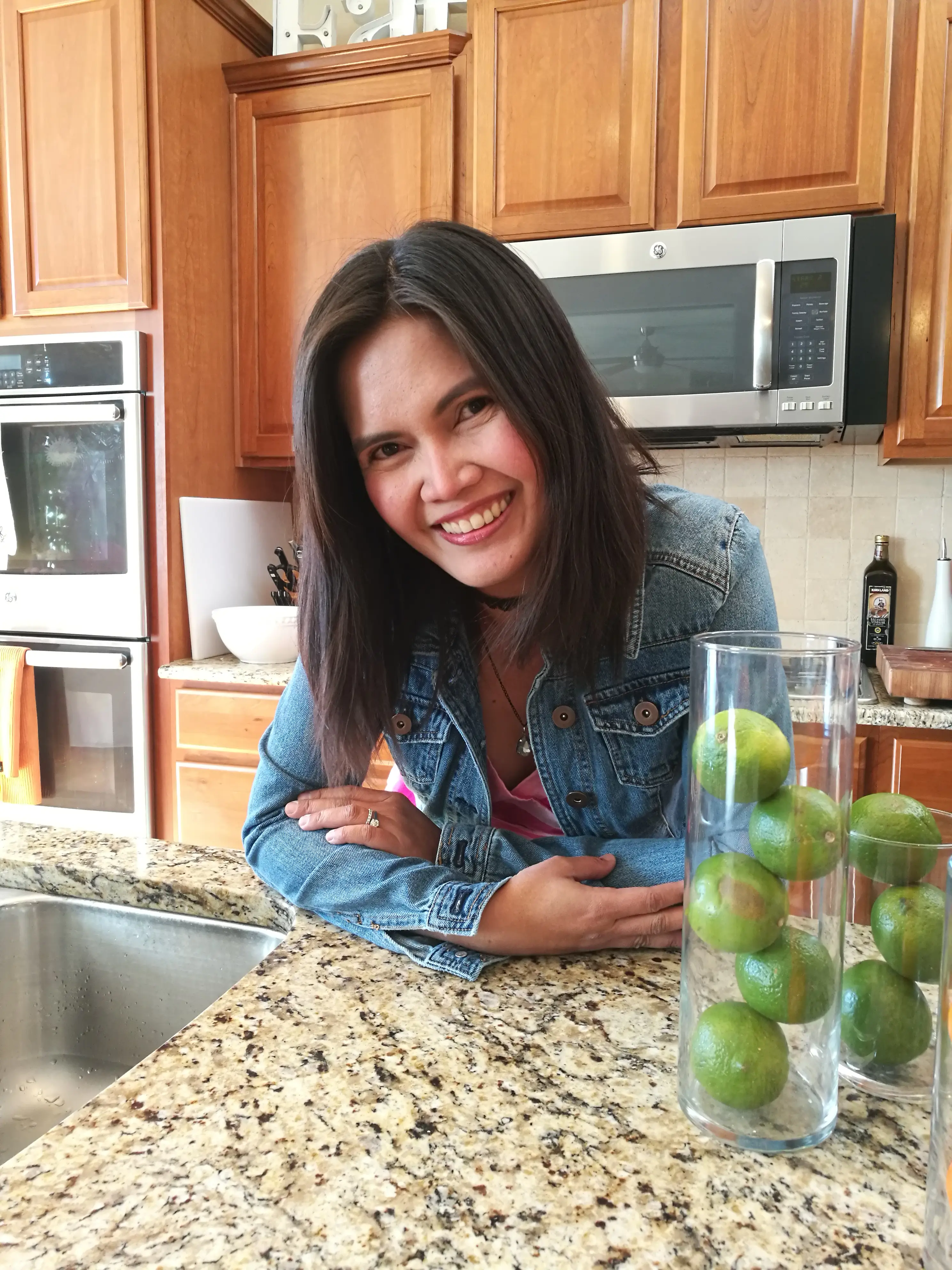 A smiling woman in a denim jacket leans over a kitchen counter with limes in a glass vase behind her.