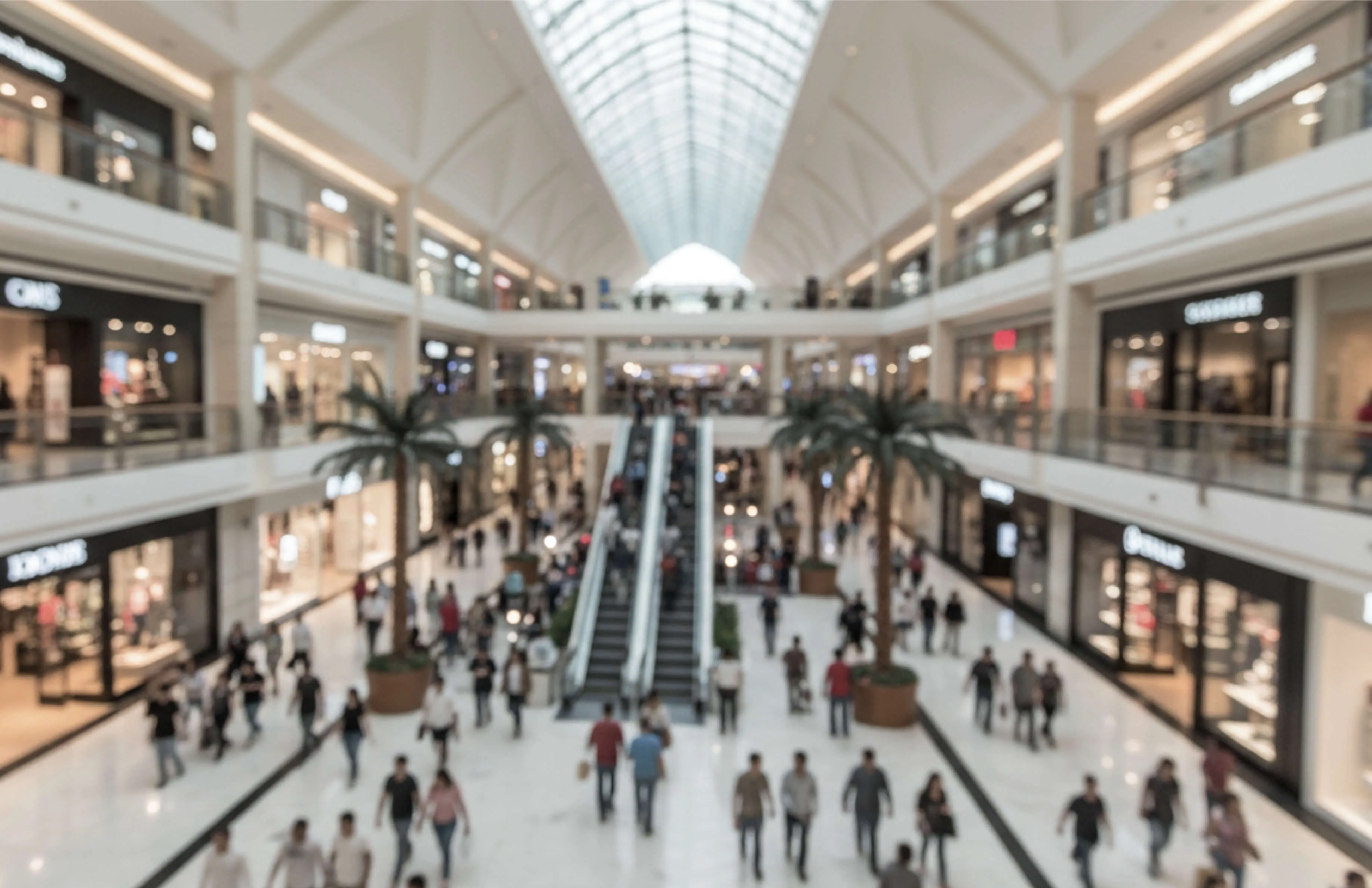 Blurred interior view of a shopping mall with people walking, escalators in the center, and shops on both sides.