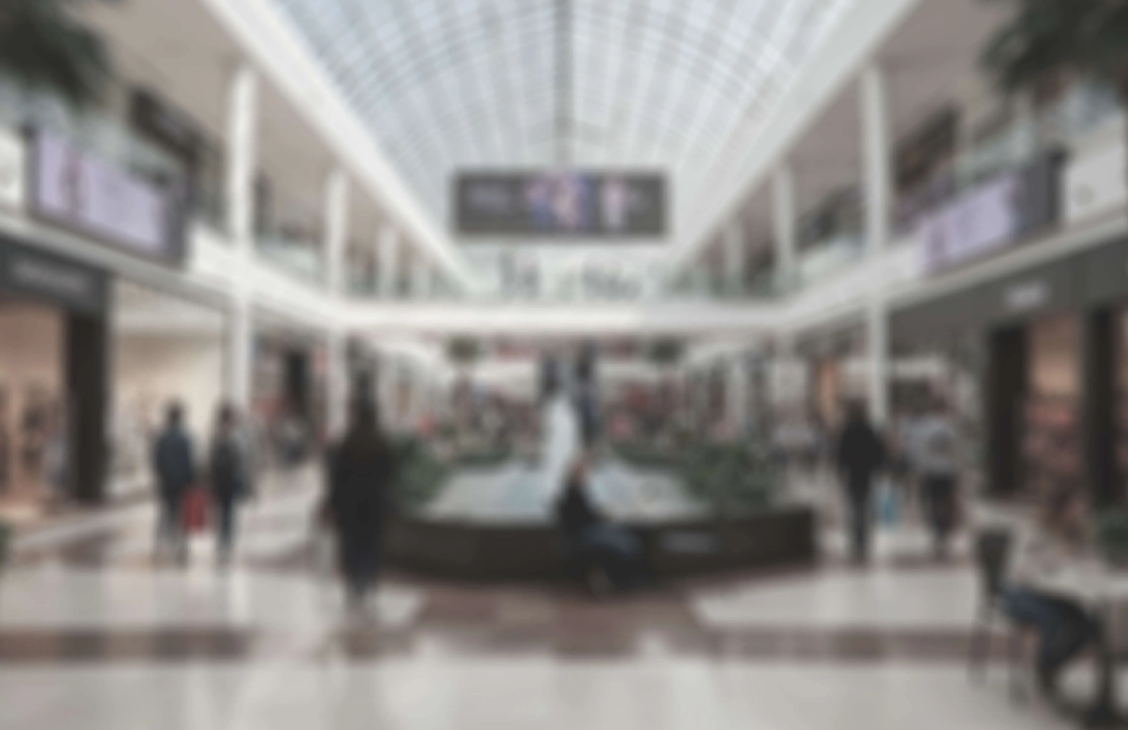 Blurry interior view of a shopping mall with people walking and sitting near a central planter.
