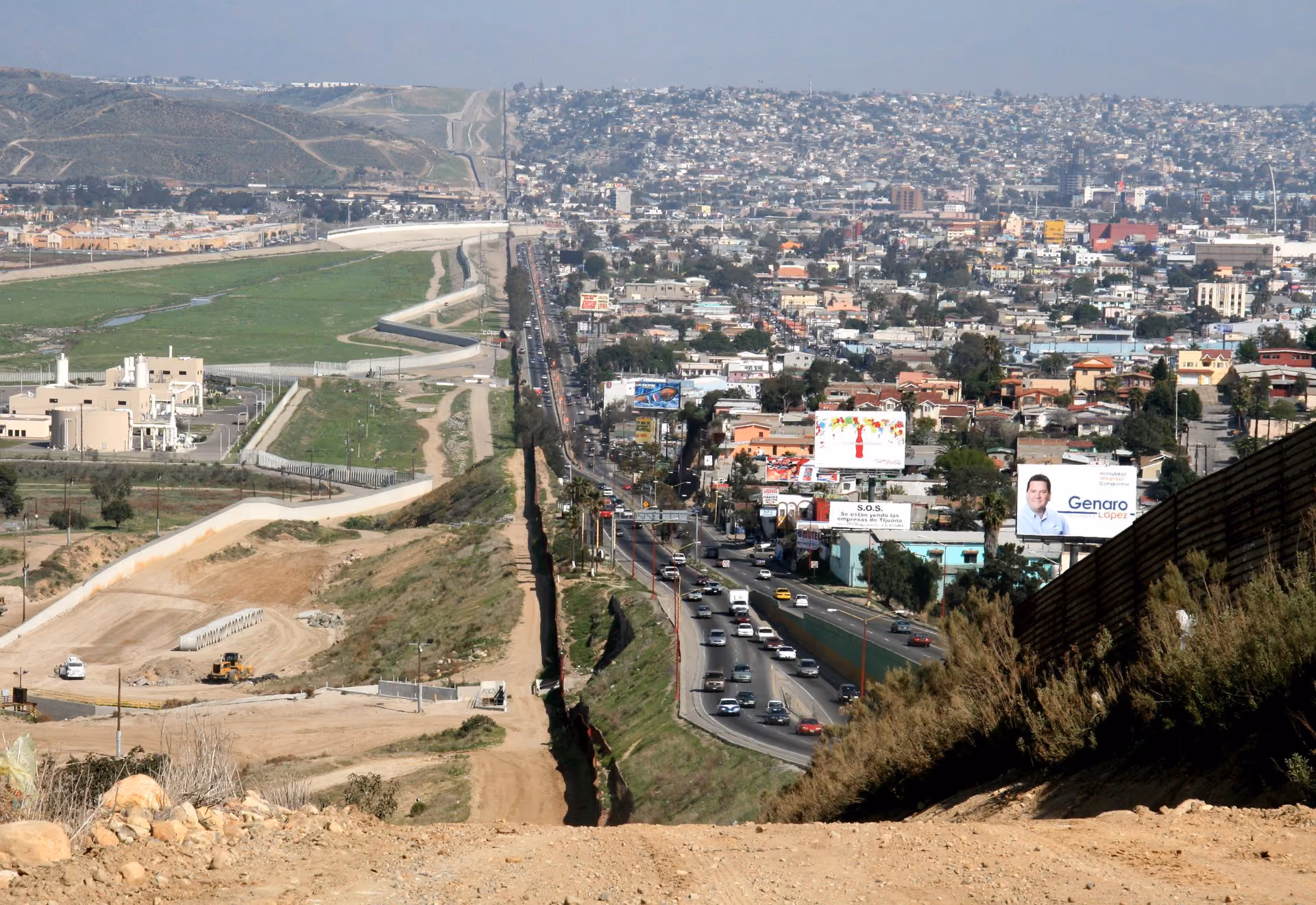 Aerial view of the Mexico-United States border wall separating Tijuana's dense urban buildings from rural San Diego terrain.