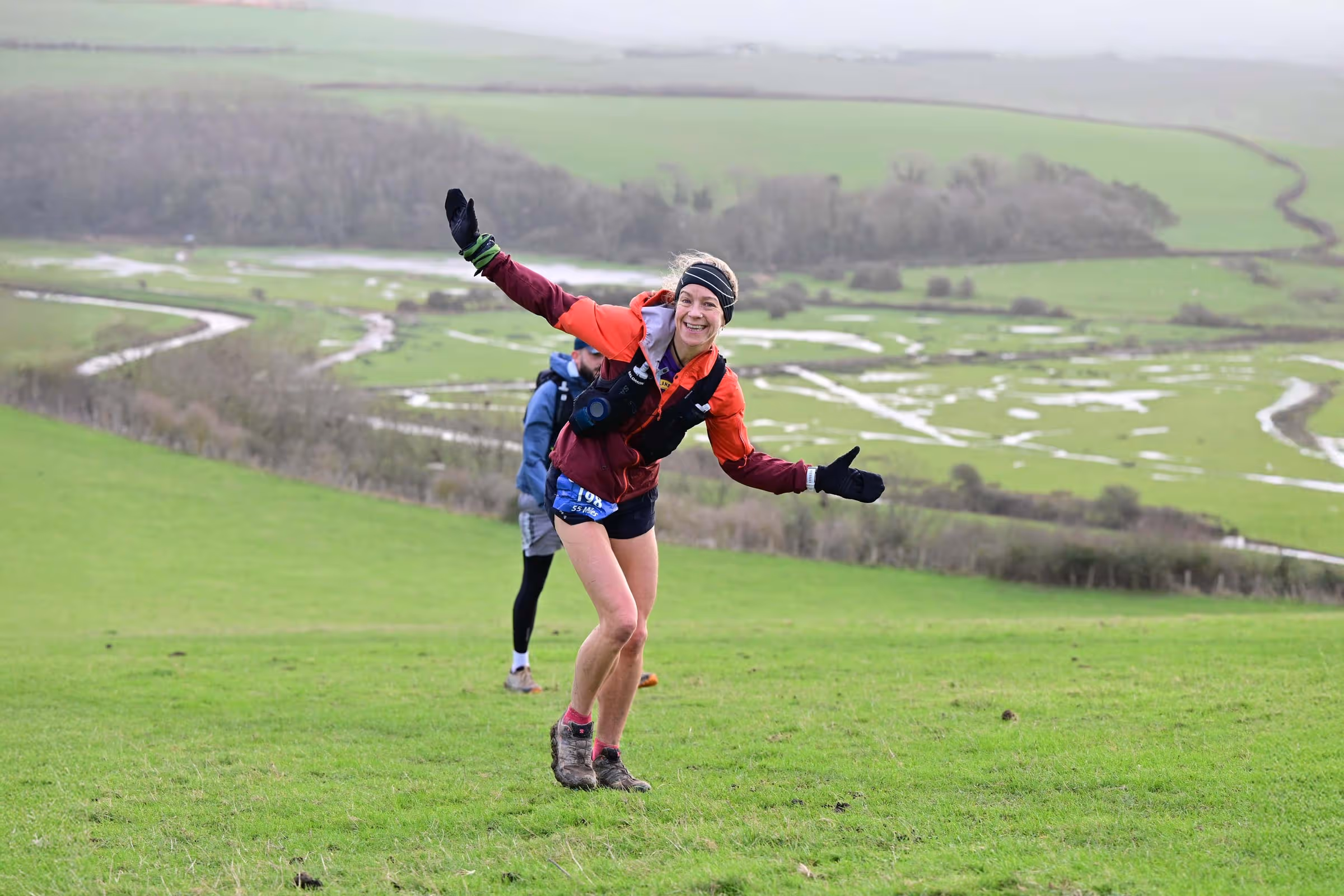 Woman running on a grassy hill, smiling and extending her arms, with another runner behind her.