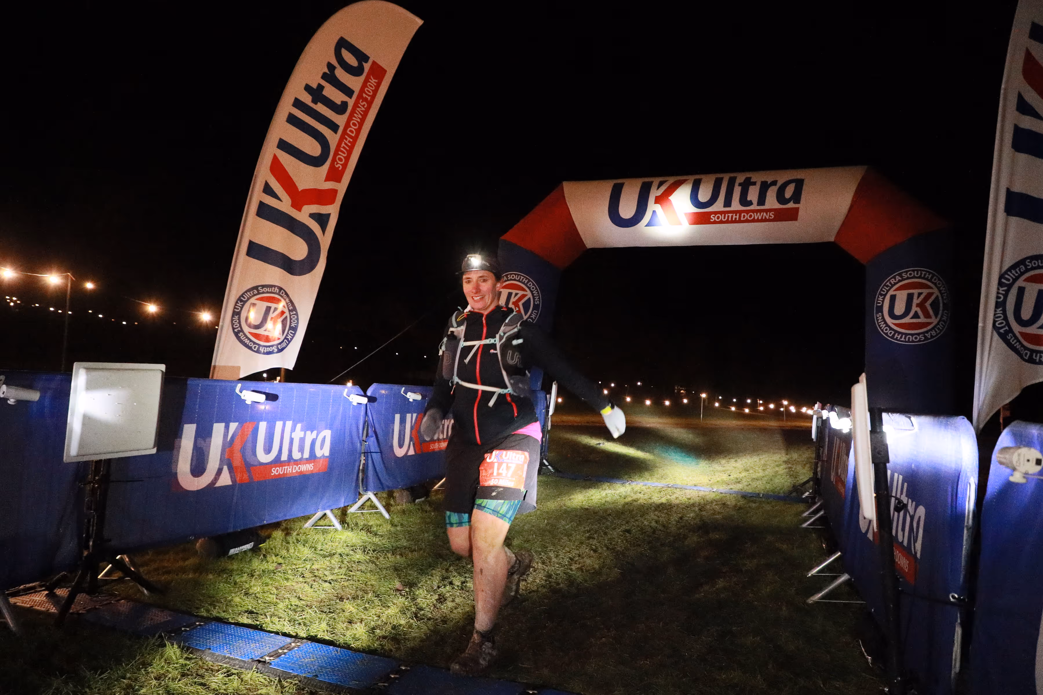 Runner finishes race at night under an illuminated "UK Ultra" arch, wearing a headlamp and gear.