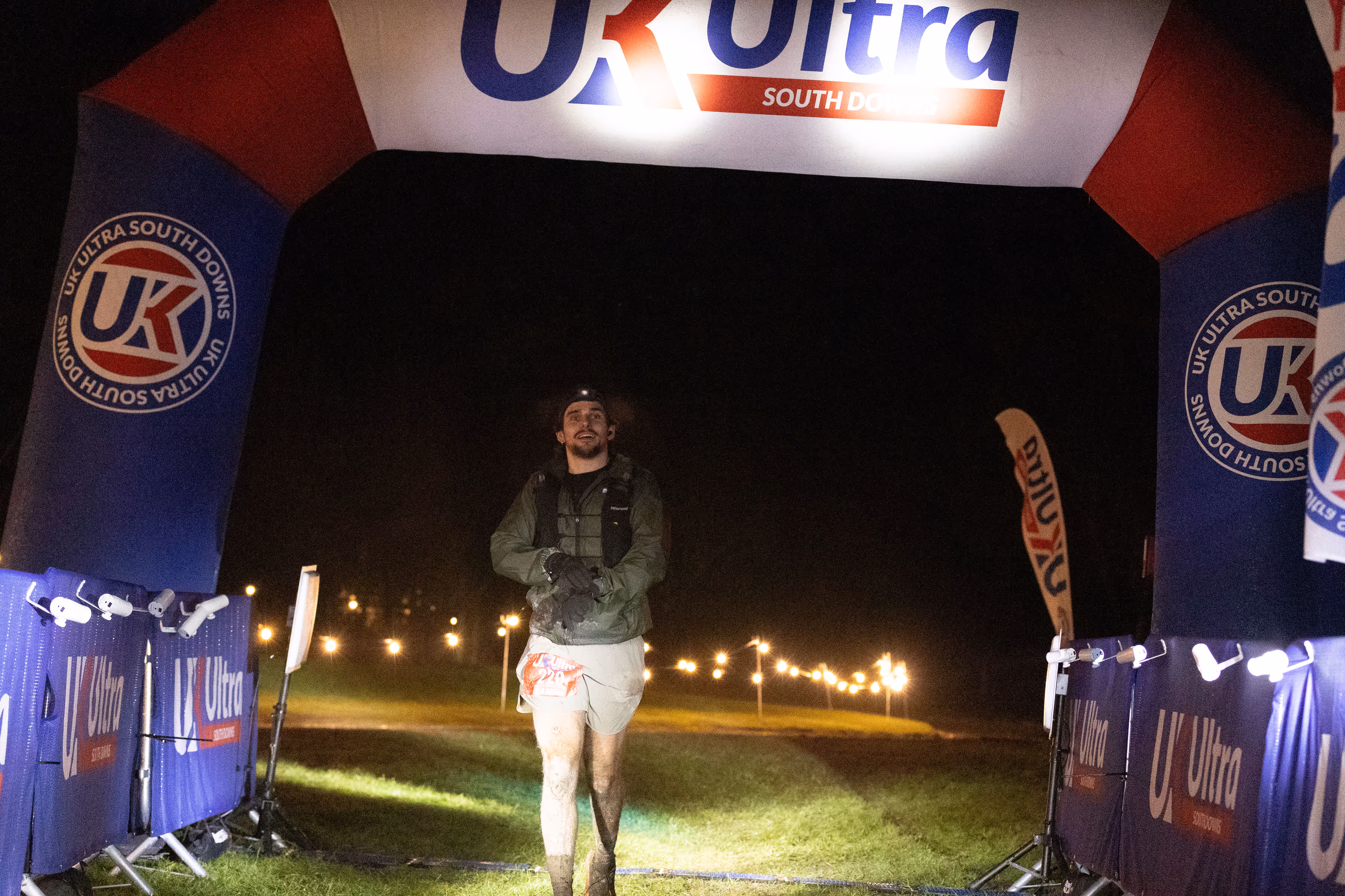 Runner in muddy shorts crossing night-time race finish line with UK Ultra banner overhead.