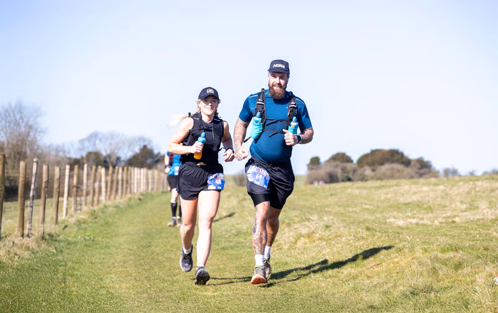 Man and woman running together on a grassy path, both wearing athletic gear and holding water bottles.