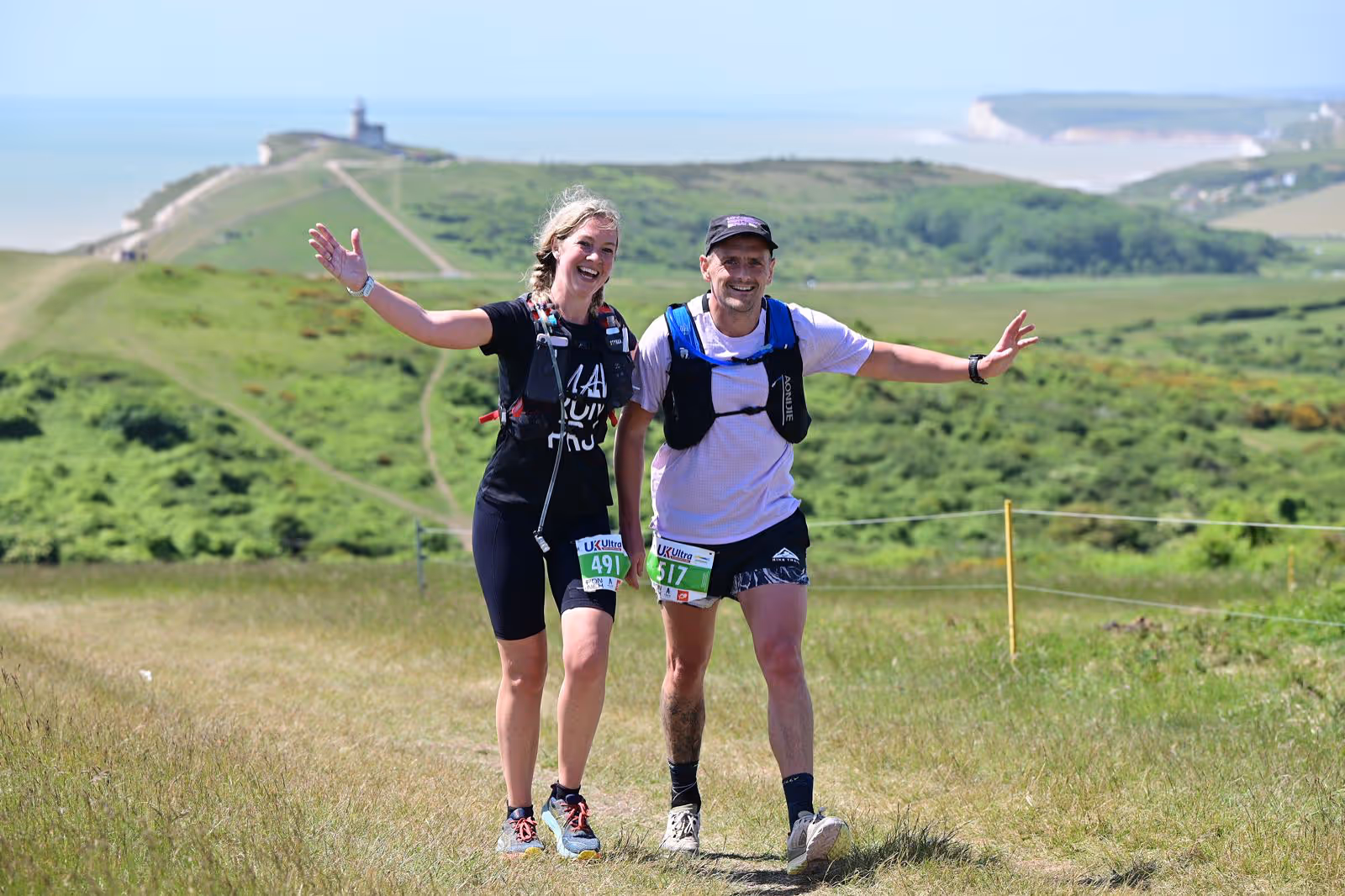 Two runners smiling and waving while walking on a grassy trail with scenic views in the background.