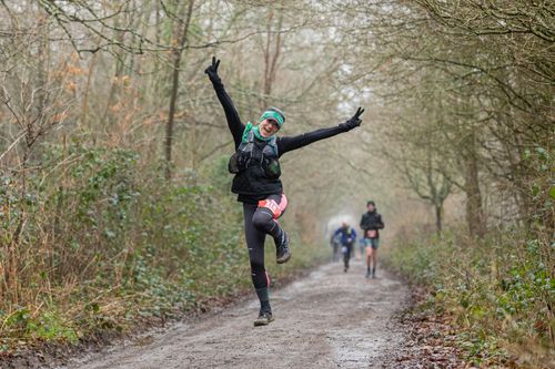 Person joyfully jumps on a muddy forest trail, while other runners approach from behind on a winter day.