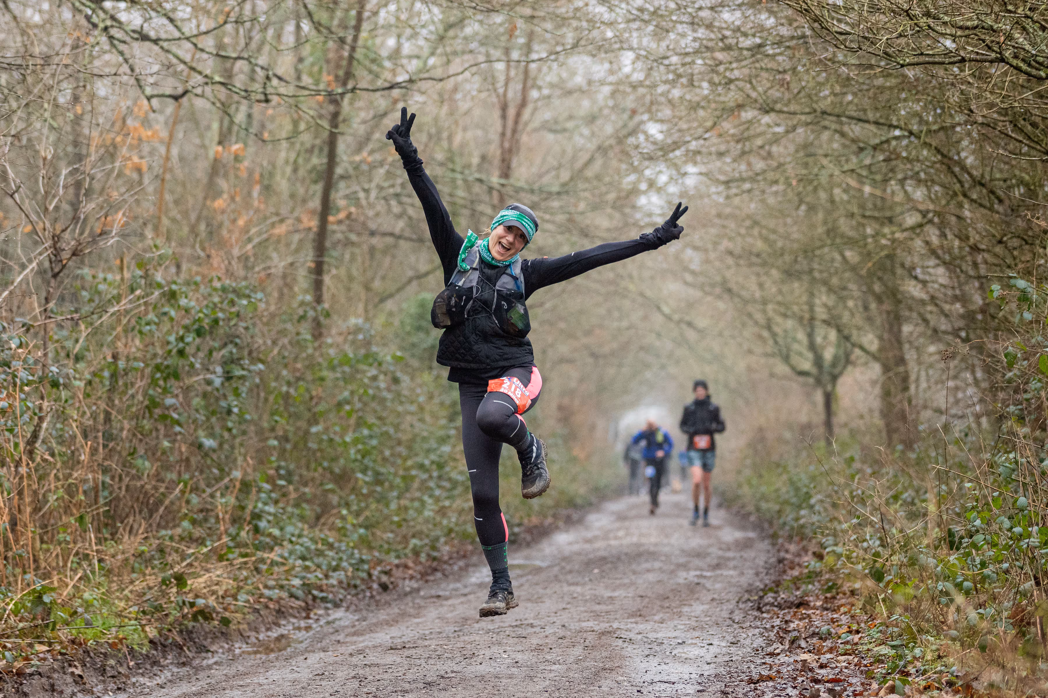 Person joyfully jumps on a muddy forest trail, while other runners approach from behind on a winter day.