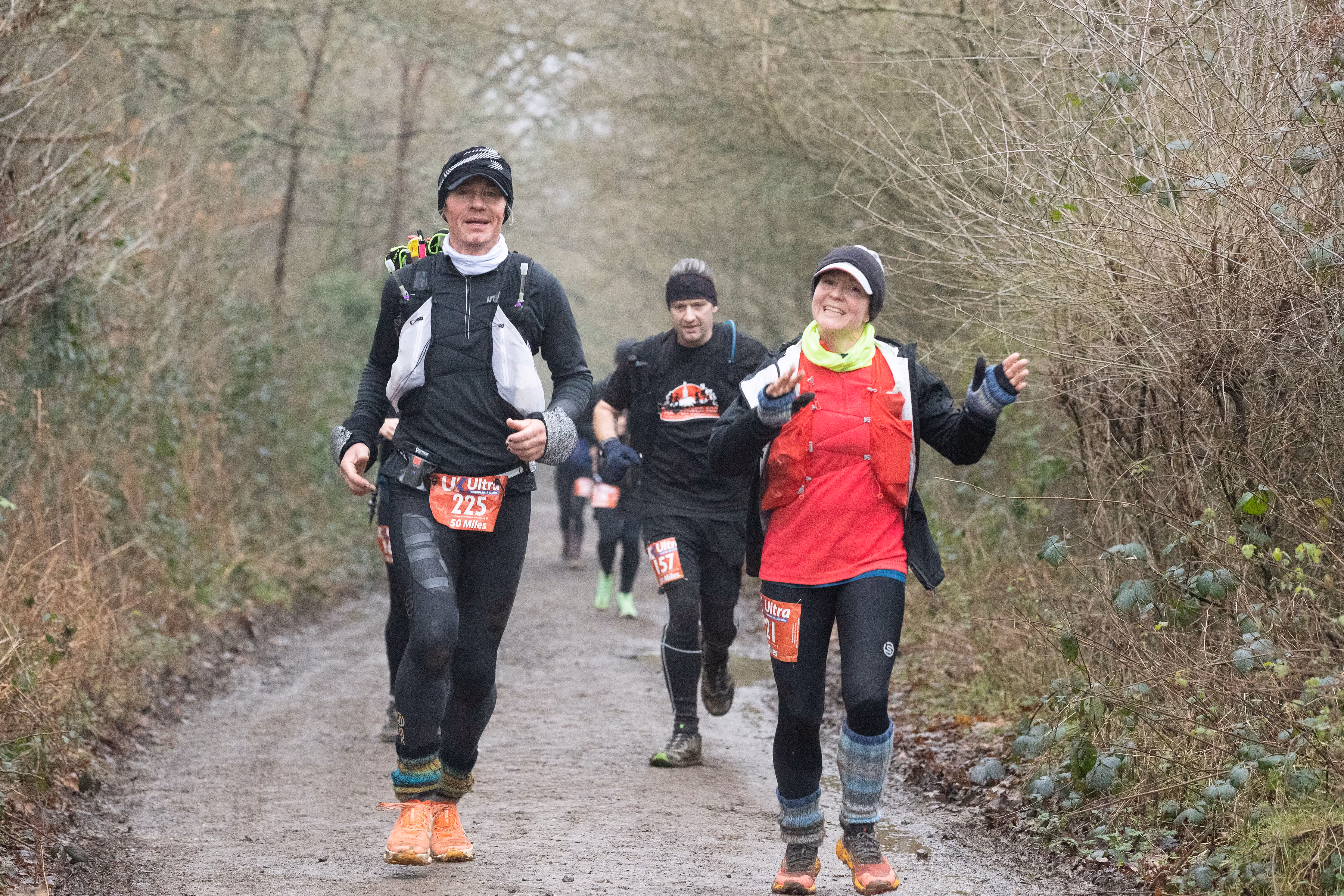 Two people in athletic gear running on a forest trail, smiling and wearing race bibs.