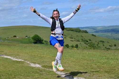Person in athletic gear jumping with arms raised, smiling on a grassy landscape with hills in the background.