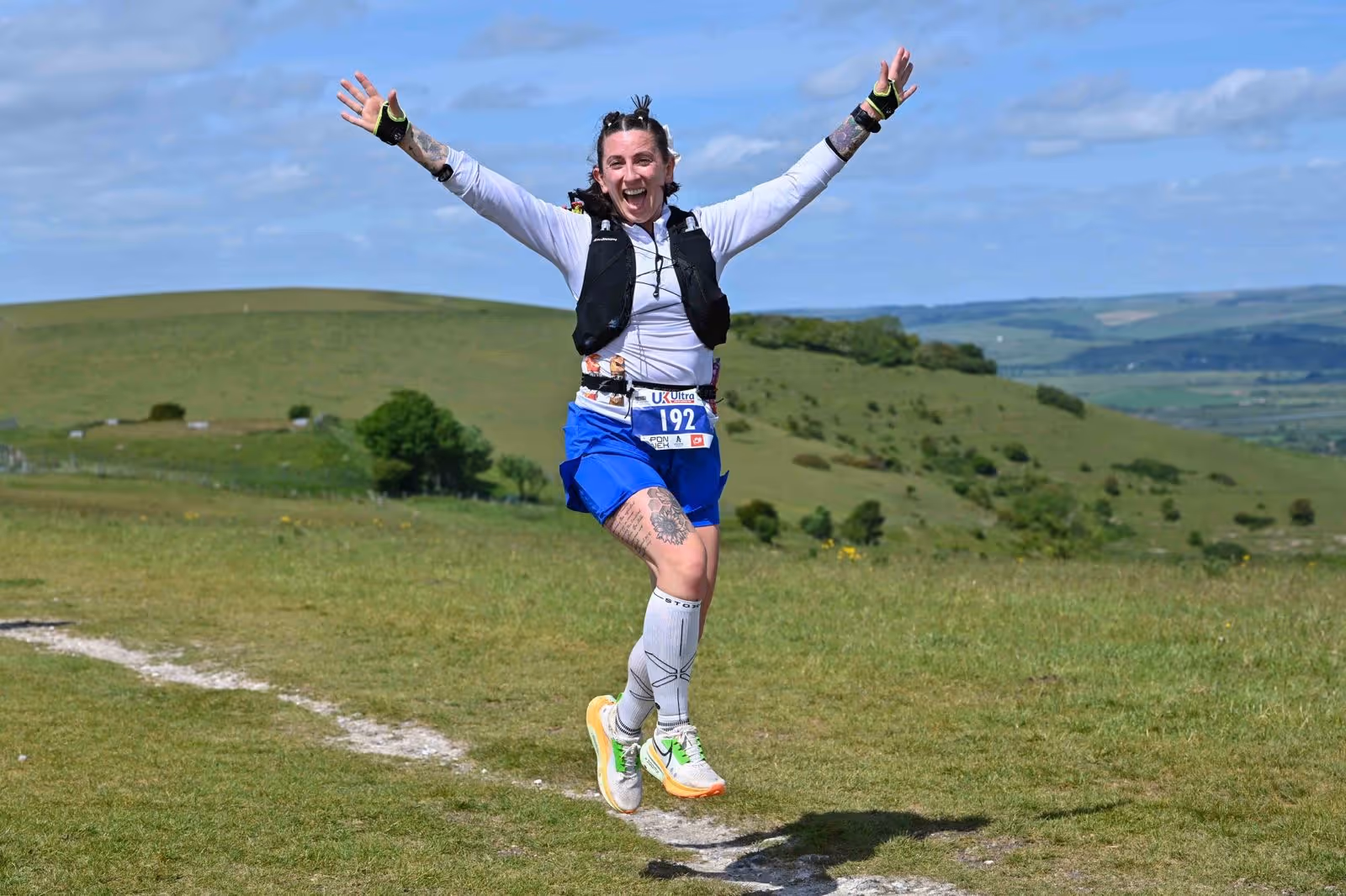 Person in athletic gear jumping with arms raised, smiling on a grassy landscape with hills in the background.