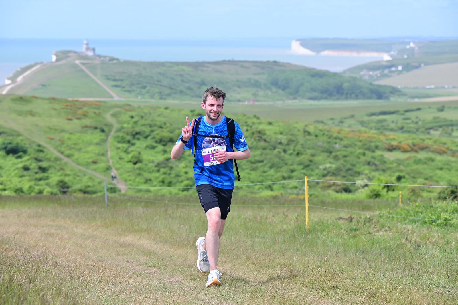 A person runs on a grassy hill, smiling and holding up two fingers, with hills in the background.