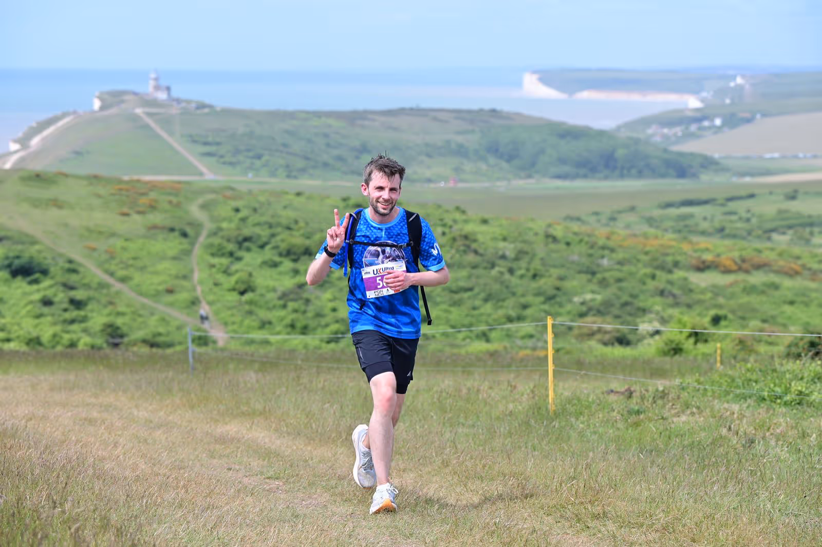 A person runs on a grassy hill, smiling and holding up two fingers, with hills in the background.