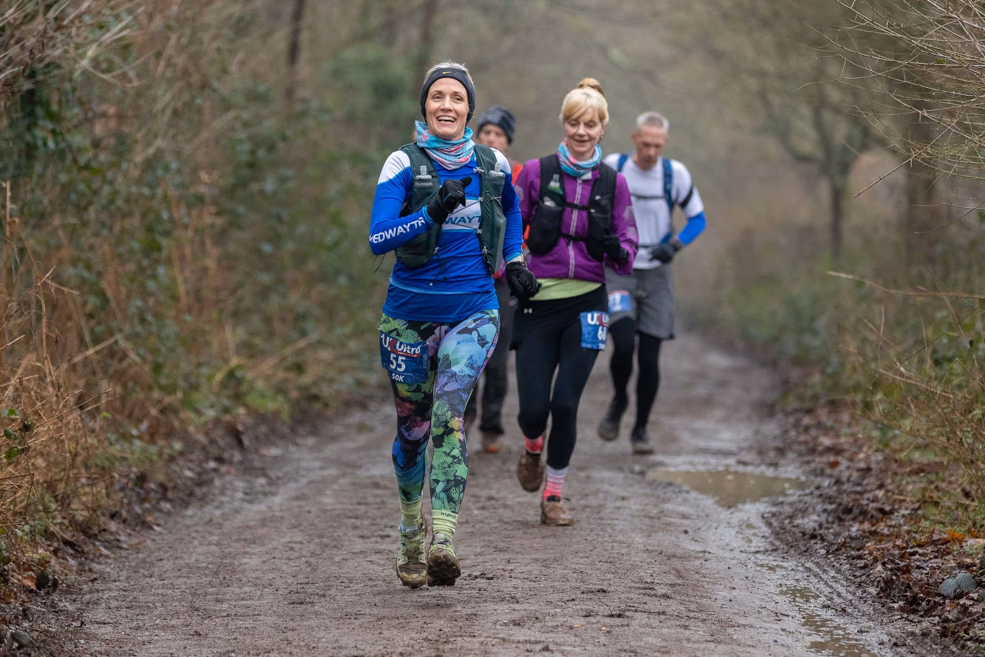 Four people running on a muddy path in a forest during a race.