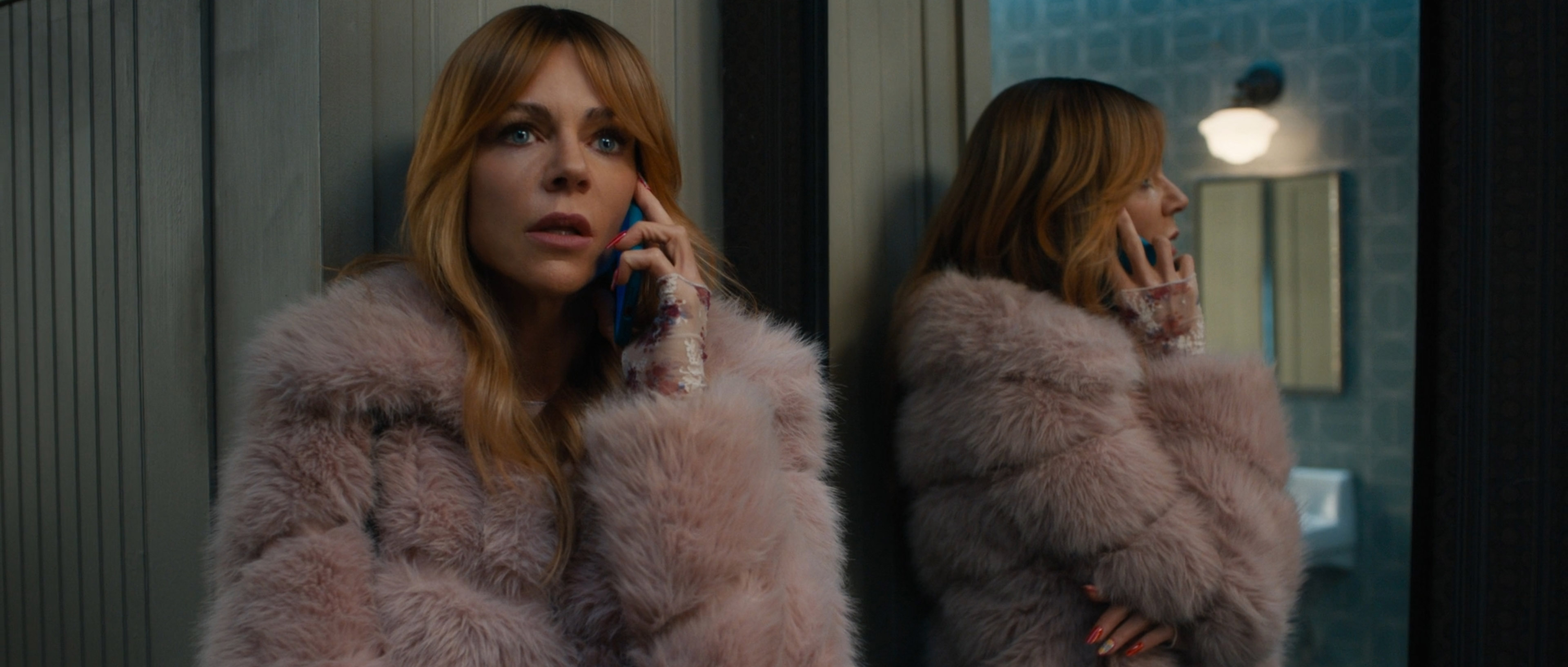 Woman with long blonde hair wearing a pink fur coat talking on a blue phone, reflected in a mirror.