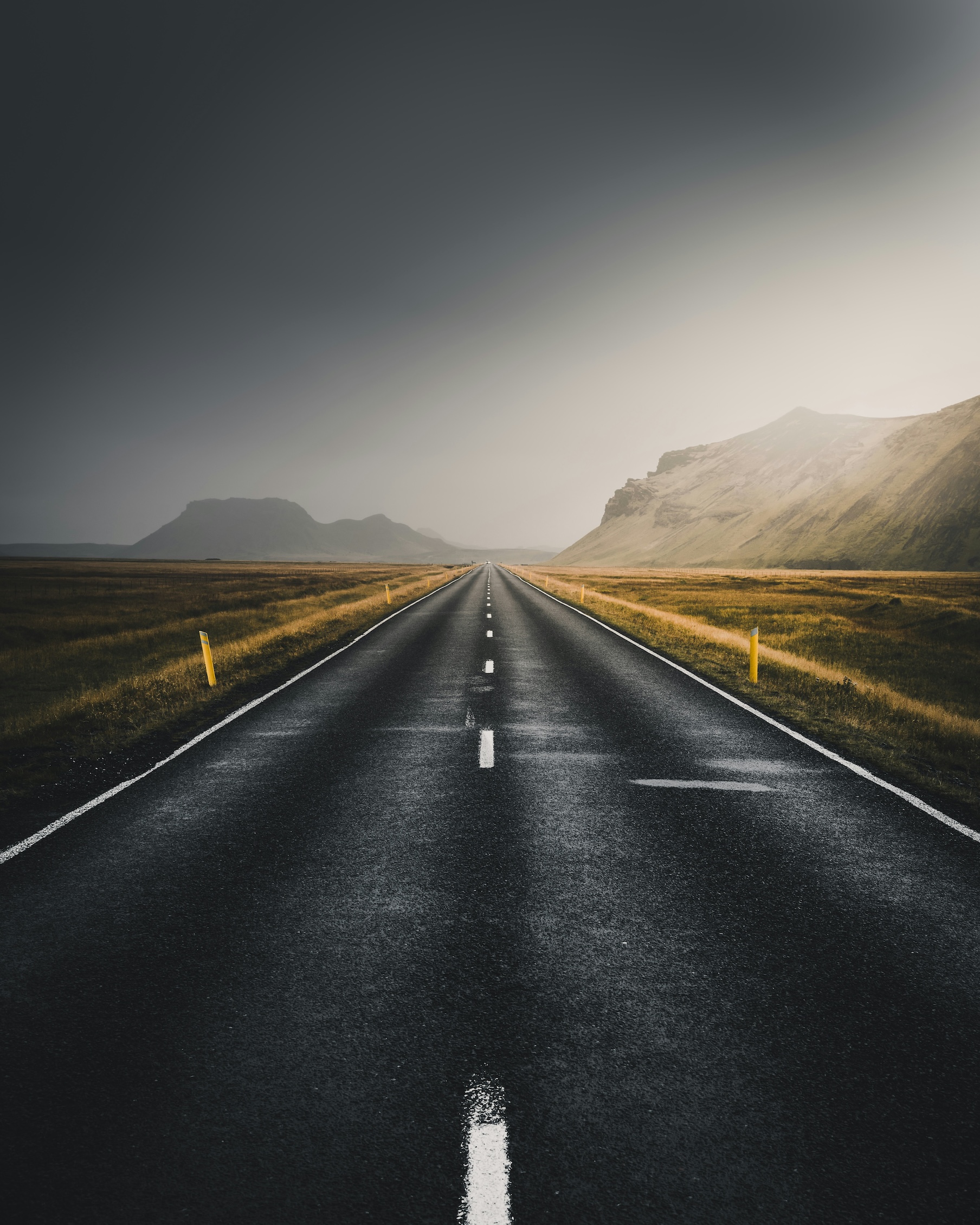 Straight empty highway stretching into the distance with mountains under a cloudy sky.