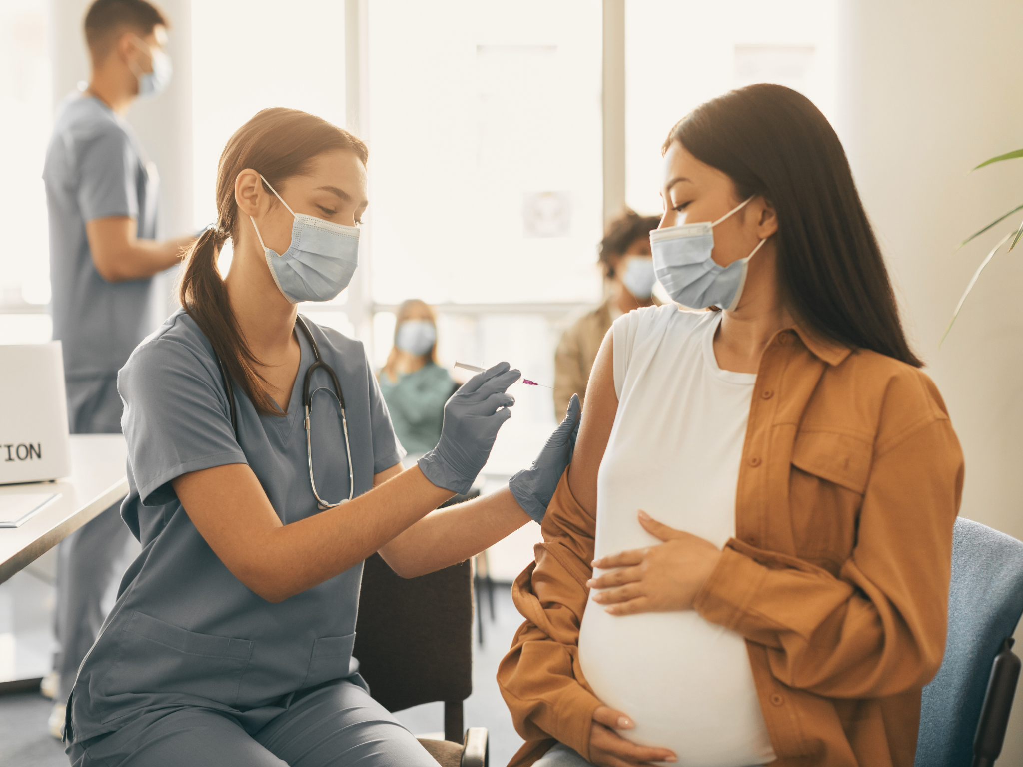 Nurse in light blue scrubs and blue gloves vaccinates a pregnant woman in a white shirt. Both are wearing medical masks. 