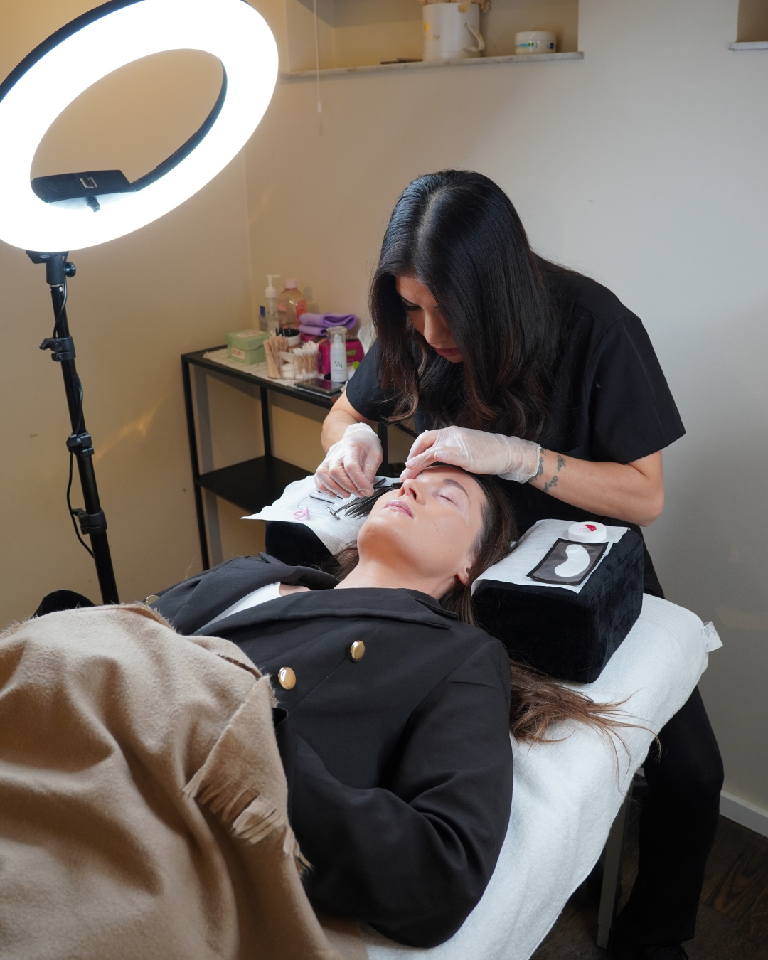 Beautician wearing gloves performing eyelash extension on a client lying on a treatment bed.