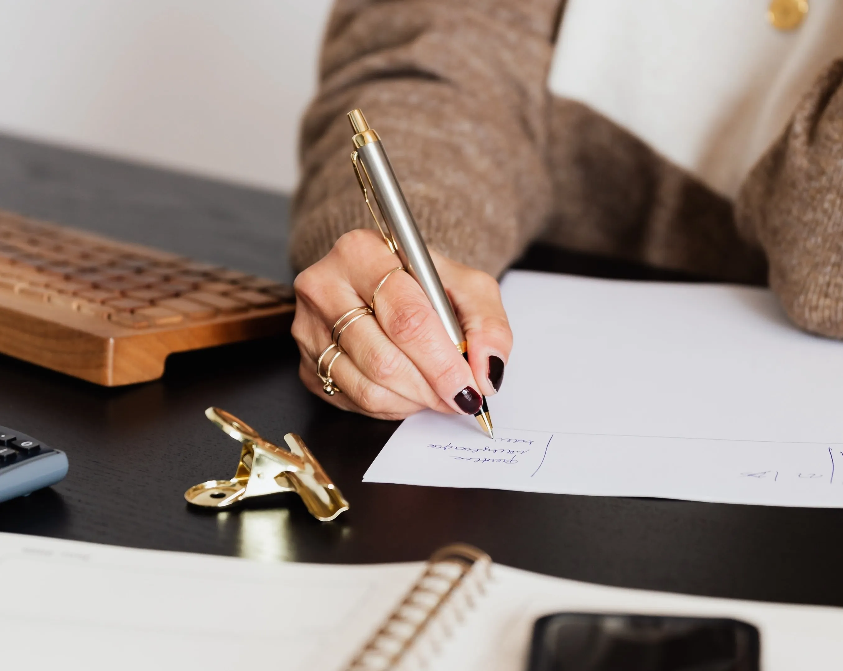 Hand writing with gold pen on paper, near calculator and wooden keyboard