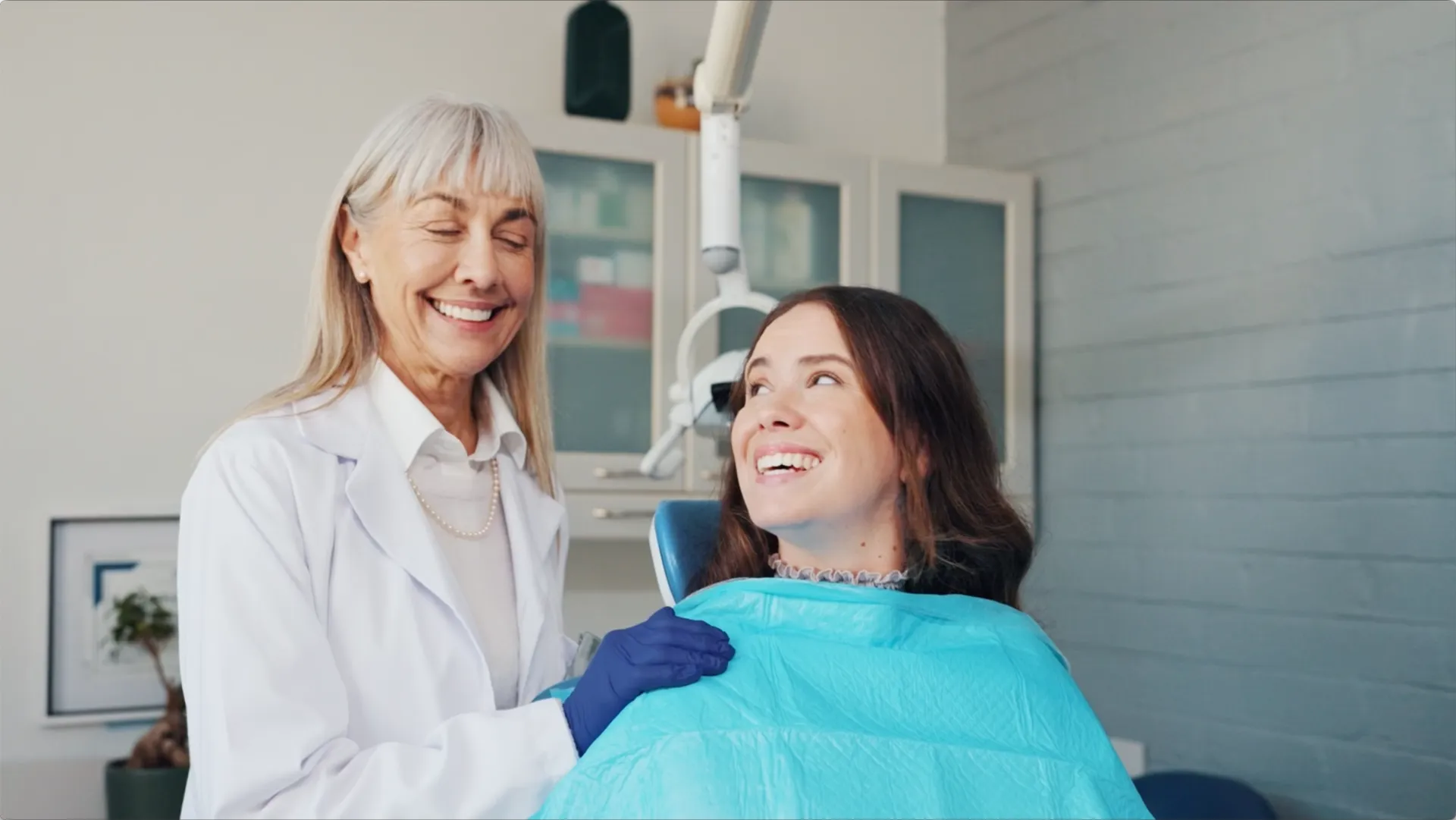 Smiling female dentist standing next to a happy female patient seated in a dental chair with a blue bib.