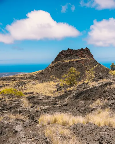 Volcanic rocky hill with sparse vegetation under a bright blue sky with fluffy white clouds.