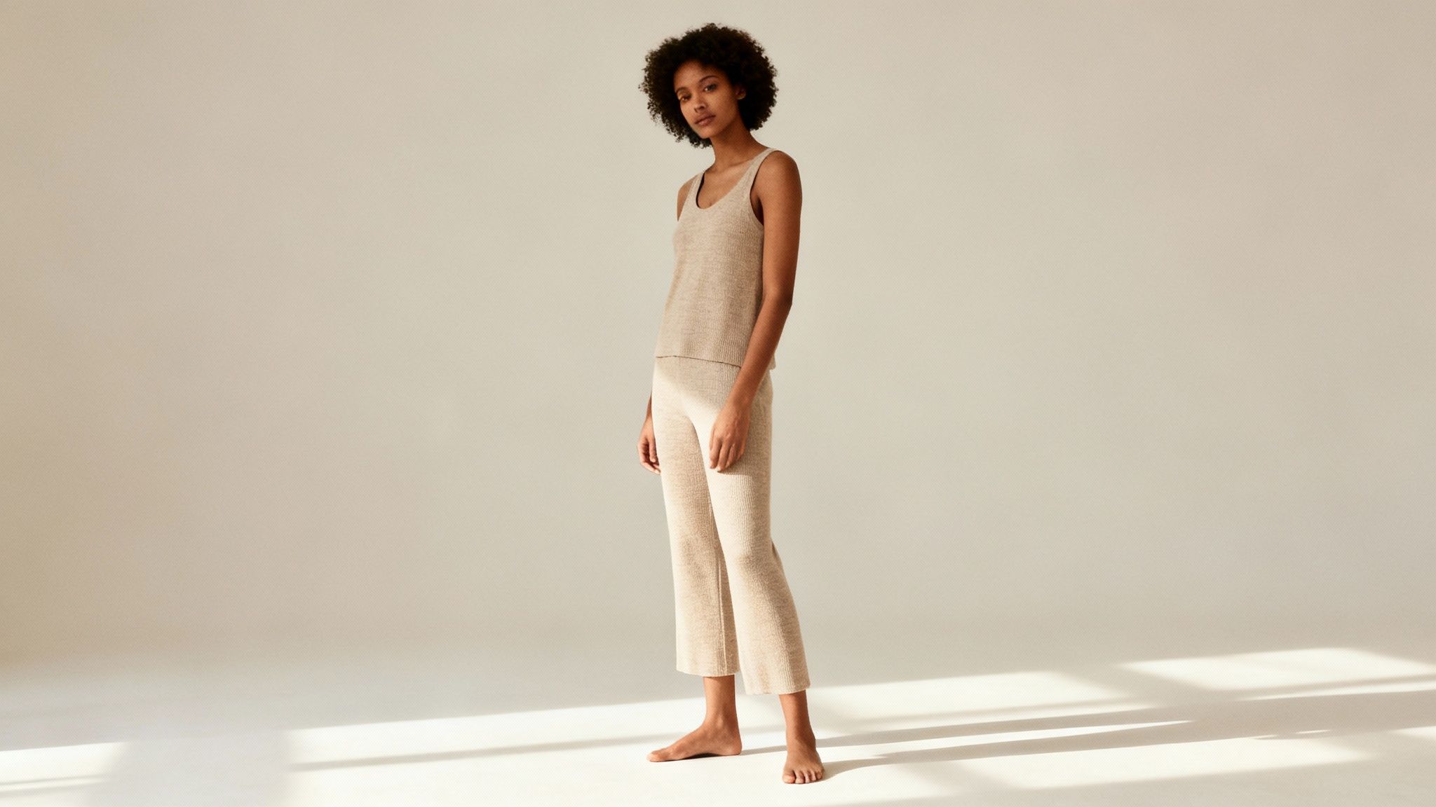 A young woman with curly hair wearing a beige knit tank top and matching pants barefoot in a studio.