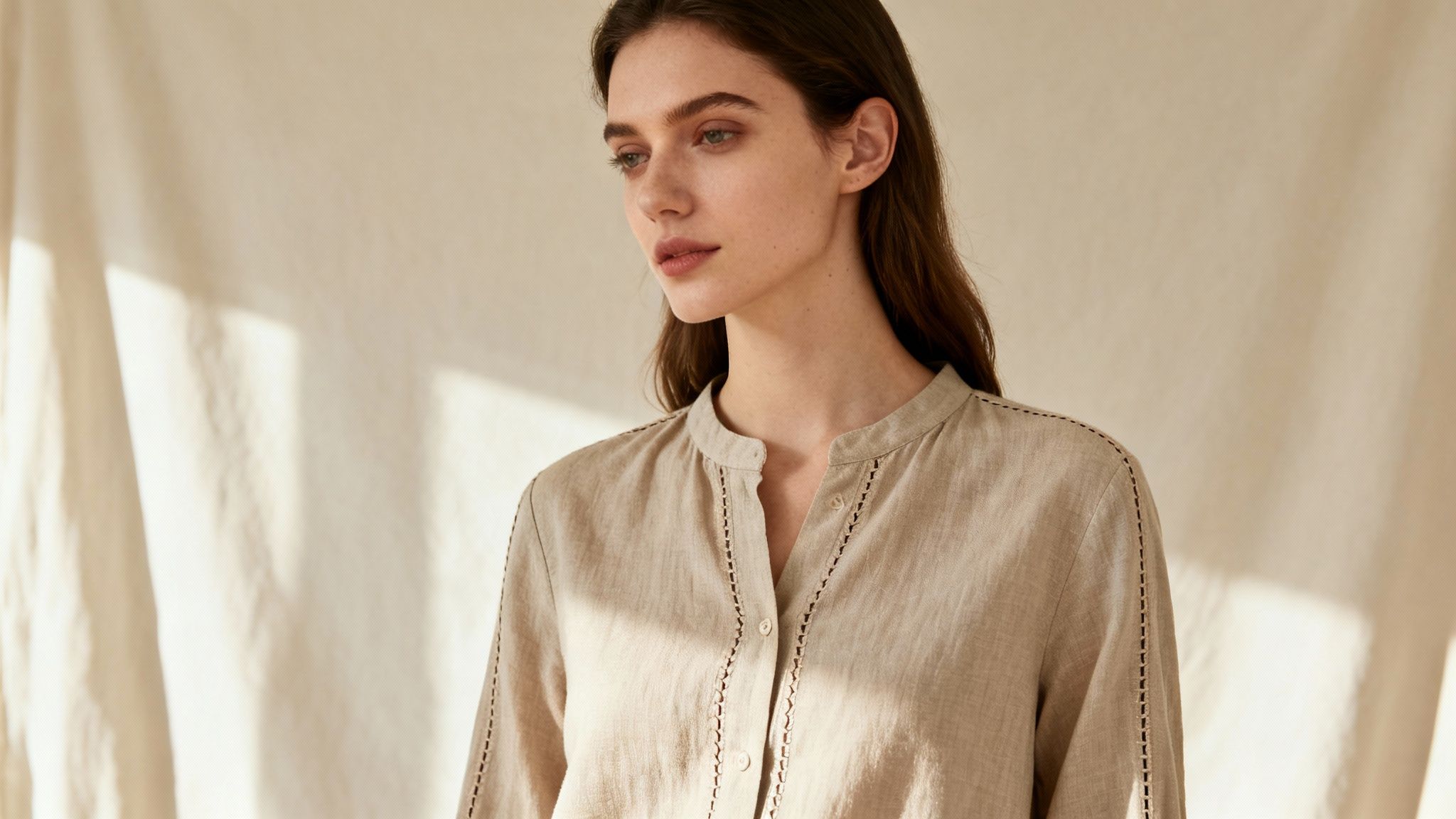 A young woman with long dark hair wears a beige linen shirt with delicate stitching, looking thoughtfully to the side.
