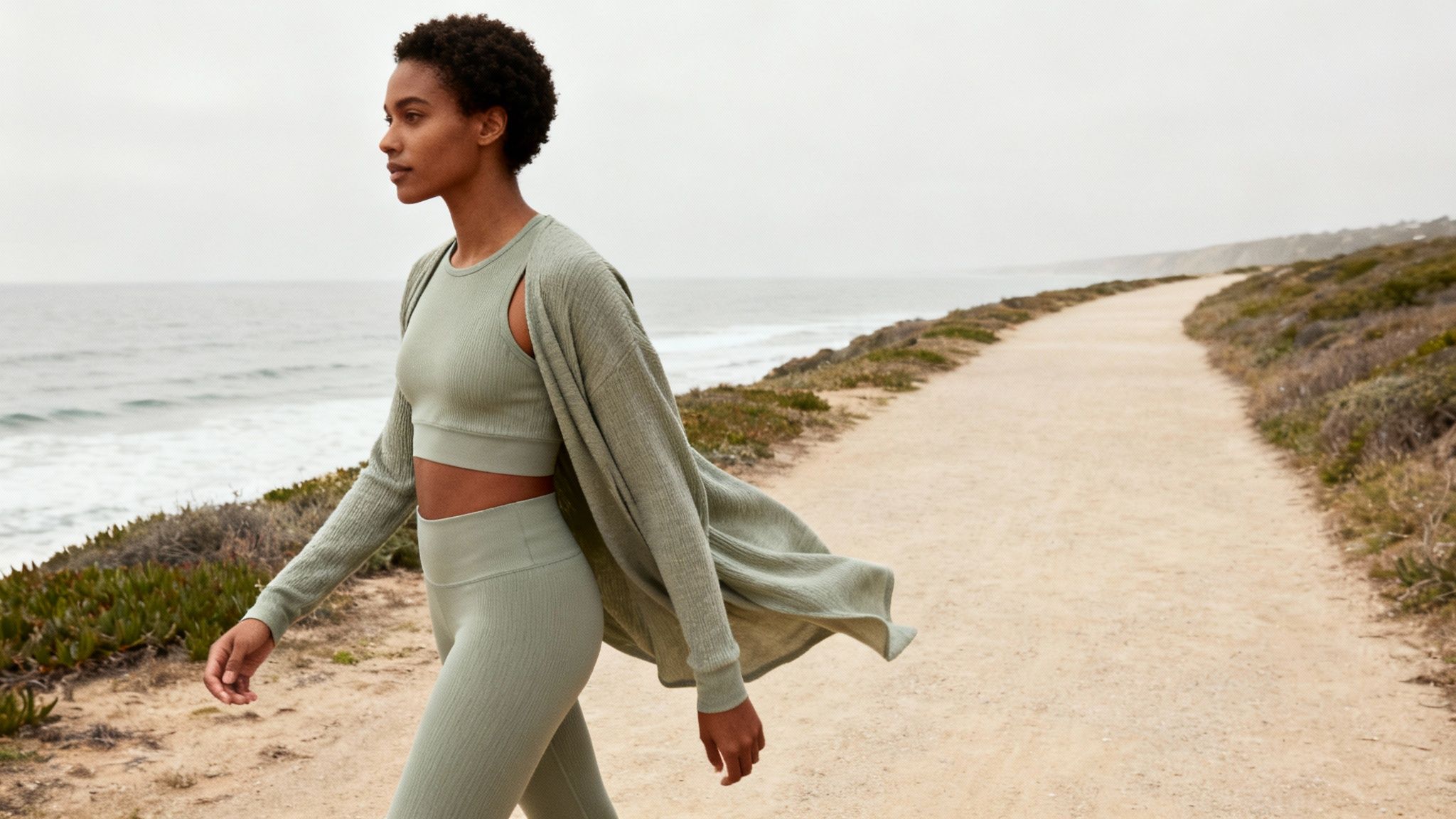 A woman in a green ribbed activewear set walks on a coastal path overlooking the ocean.