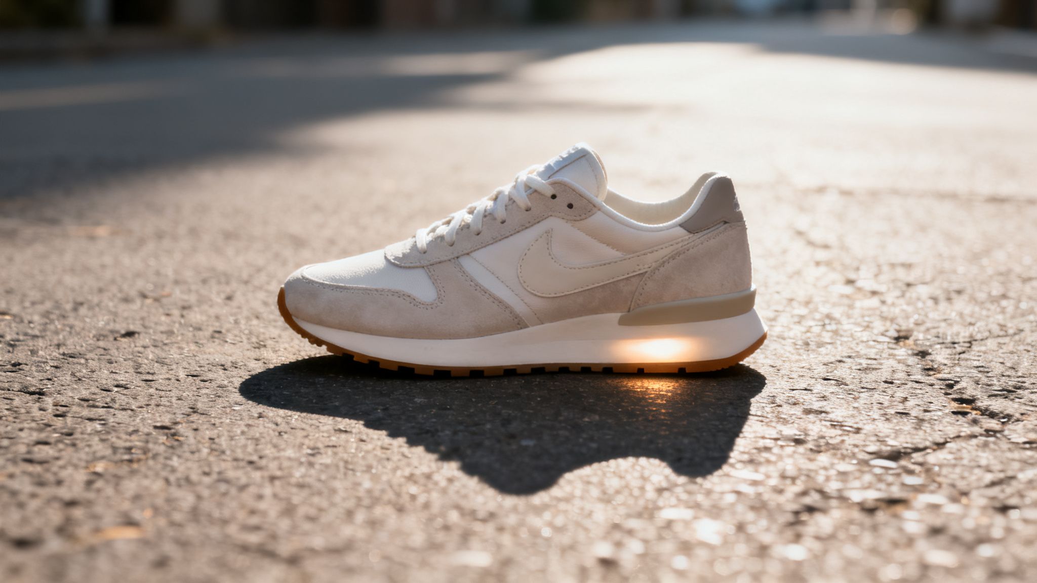 A cream and beige Nike sneaker with a glowing sole sits on a sunny asphalt road.