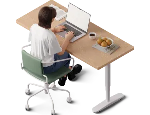 Person with short dark hair sitting on a green office chair working on a laptop at a wooden desk with a bowl of fruit and coffee cup.