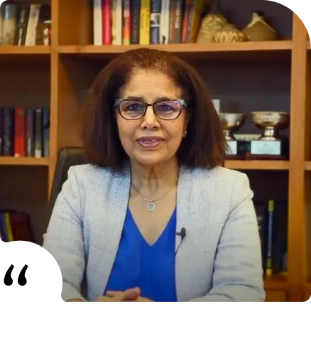 Woman with glasses and curly hair sitting at a desk wearing a light gray blazer over a blue top, with bookshelves and trophies in the background.