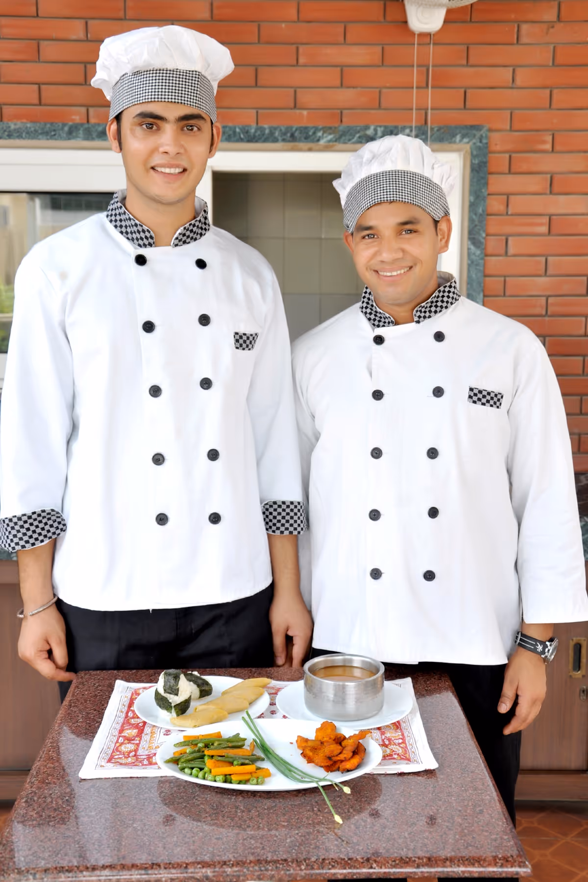 Two chefs in white uniforms and checkered hats standing behind a table with plated dishes including vegetables, fried items, and a bowl of sauce.