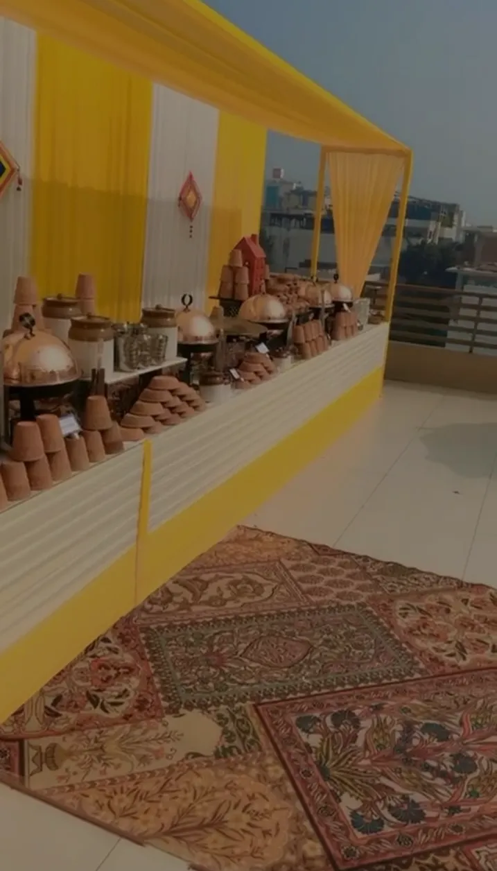 Outdoor buffet setup with traditional clay pots and copper serving dishes arranged on a yellow and white decorated table under a yellow canopy with patterned rugs on the floor.