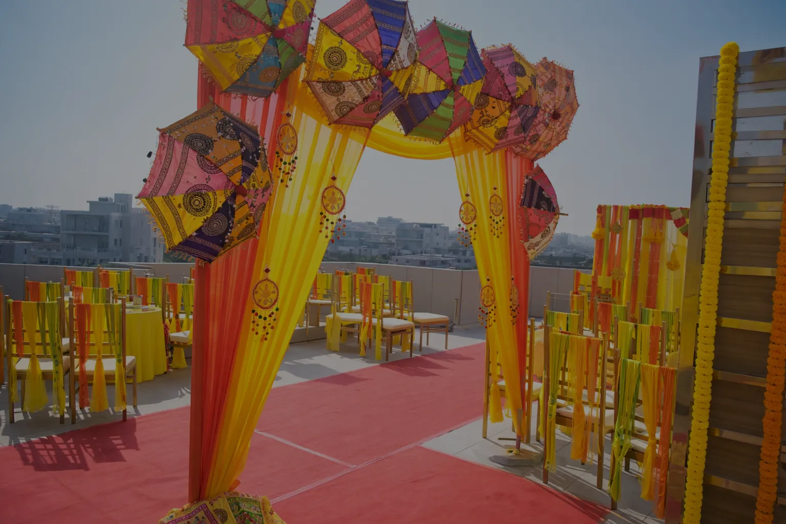 Outdoor event setup with a yellow and orange draped arch decorated with colorful umbrellas and marigold garlands with rows of chairs draped in yellow and orange fabric.