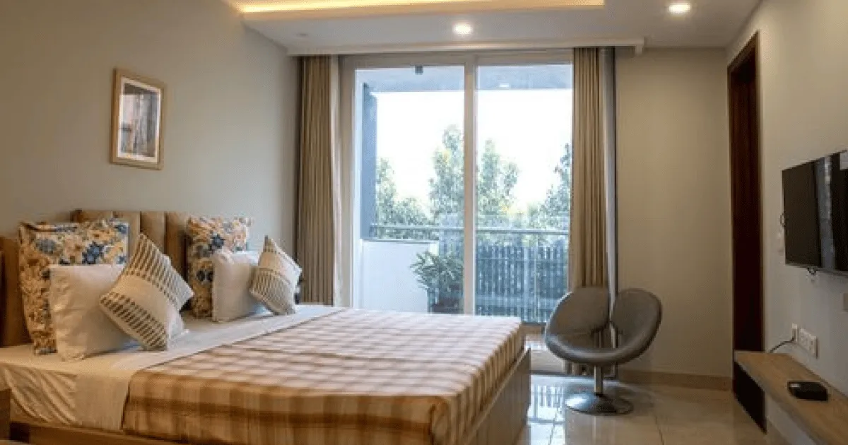 Modern bedroom with a large bed featuring plaid bedding and patterned pillows, a round chair, wall-mounted TV, and sliding glass doors leading to a balcony with plants.