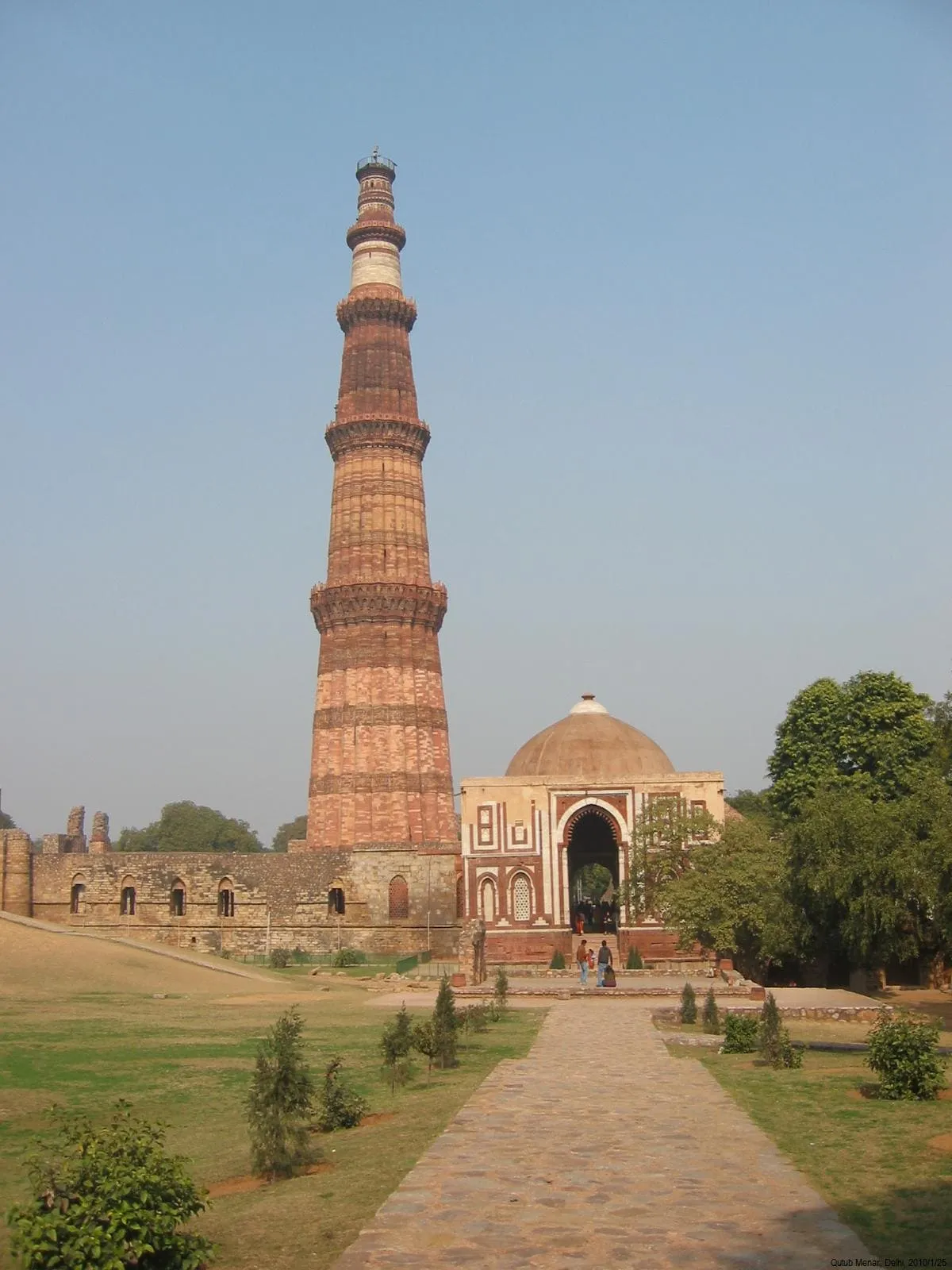 Tall historic Qutb Minar tower and adjacent ancient building with arched entrance in a park setting under clear blue sky.
