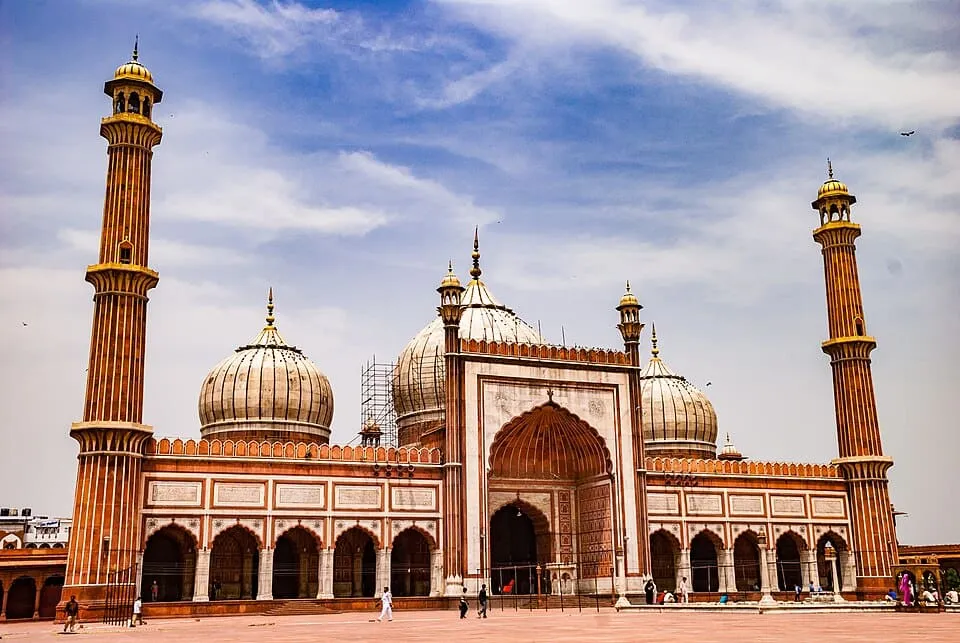 Large historic mosque with three domes and two tall minarets under a partly cloudy sky.