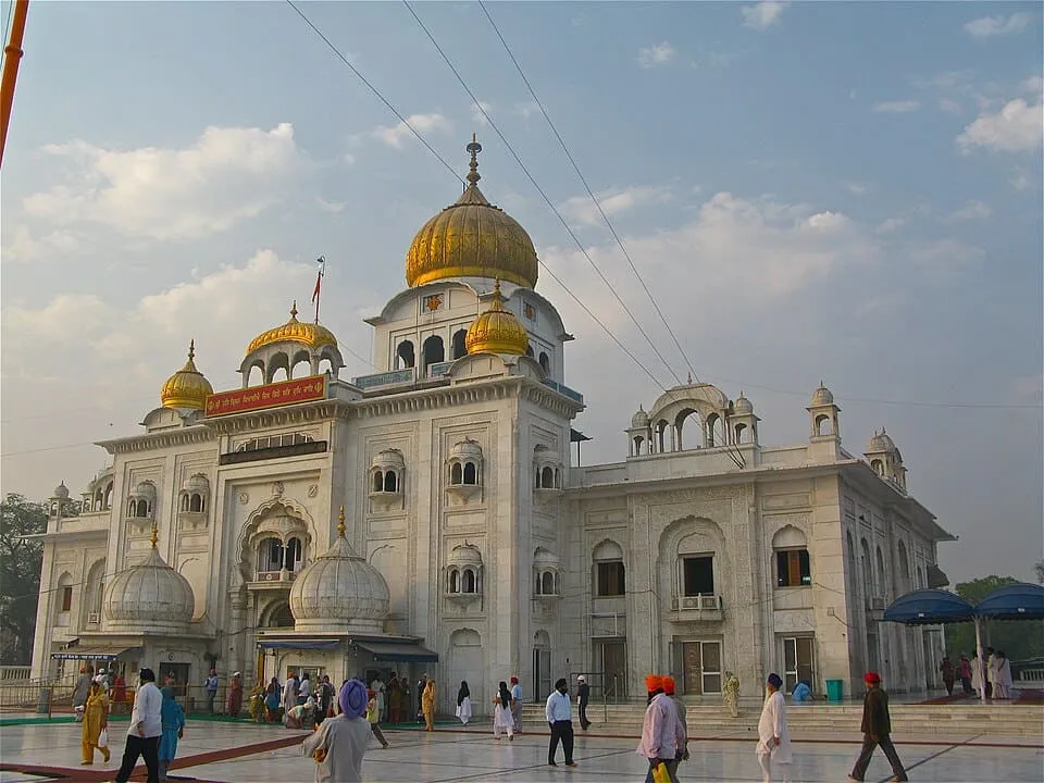 Front view of a white marble Sikh temple with golden domes under a partly cloudy sky, with visitors walking in the courtyard.