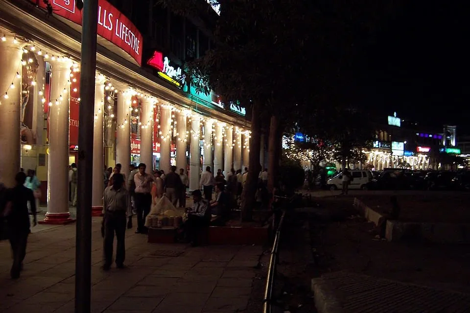 People walking and sitting near a brightly lit row of shops with white pillars at night.