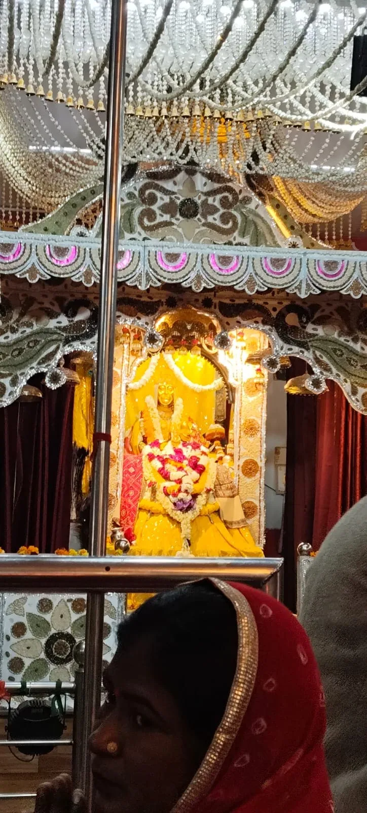 Hindu temple altar decorated with flowers, beads, and garlands featuring a golden deity statue, with a woman in a red headscarf in the foreground.