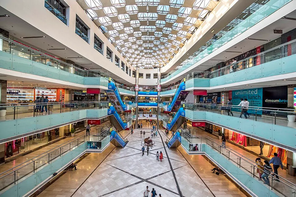 Interior of a modern multi-level shopping mall with glass railings, blue escalators, and people walking on the tiled floor under a large skylight ceiling.