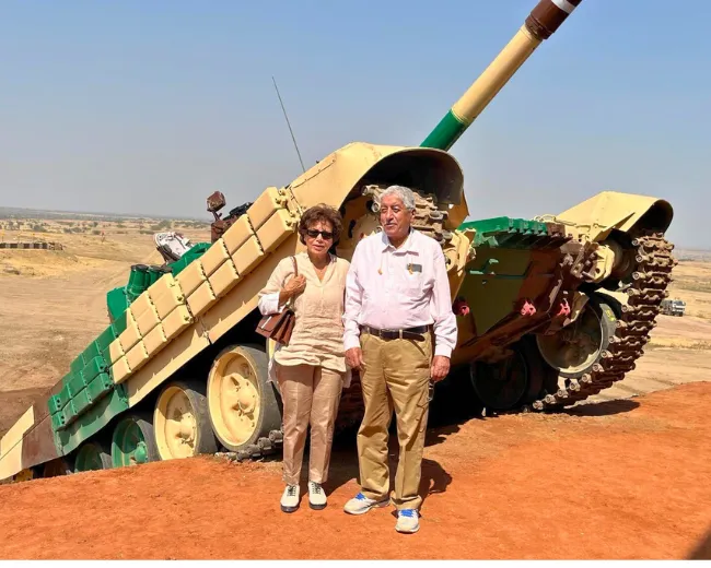 An elderly man and woman standing on reddish dirt in front of a large beige and green military tank displayed outdoors.