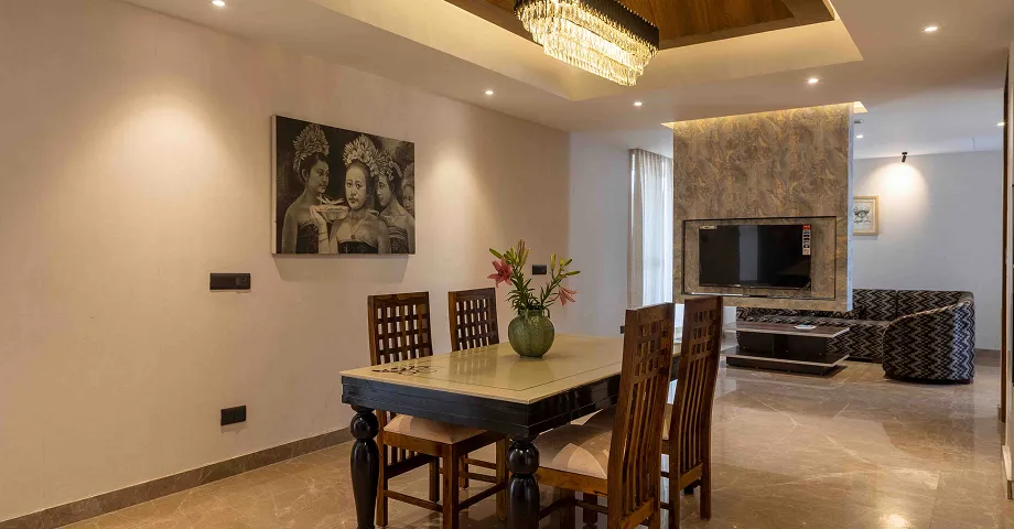 Modern dining area with wooden table and chairs, a vase with pink lilies centerpiece, beige walls, artwork of three women, and a living area with a wall-mounted TV and patterned sofa.