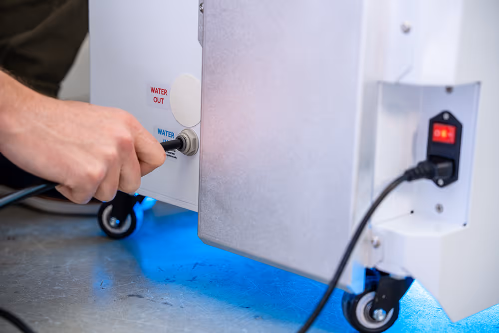 Hand plugging a water hose into a white machine labeled with water inlet and outlet ports on caster wheels.