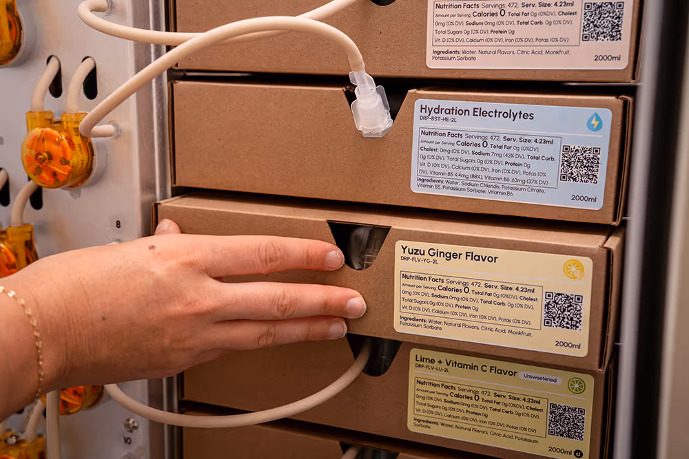 Hand accessing a boxed drink dispensing machine with labeled flavors including Hydration Electrolytes, Yuzu Ginger Flavor, and Lime + Vitamin C Flavor.