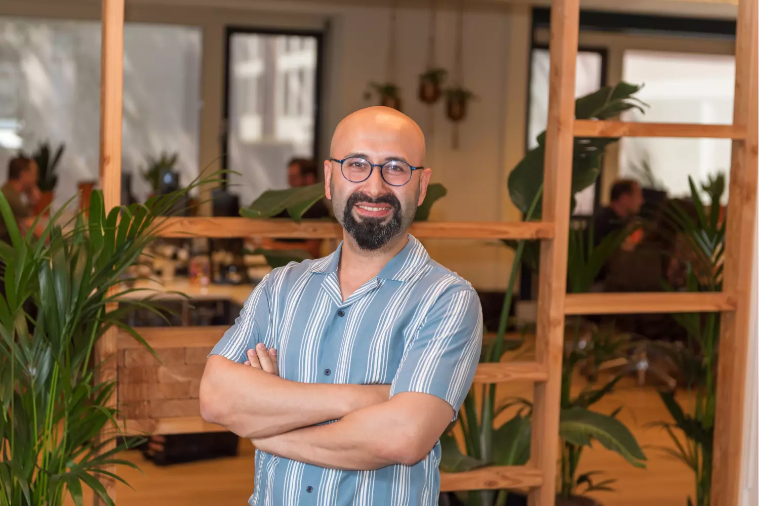 Gelukkige man met grijze baard en groenig overhemd staat in een kantoorruimte met planten achter zich.
