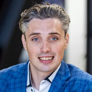 Man met licht golvend grijs haar en blauwe ogen, glimlachend in een blauw geruit jasje.