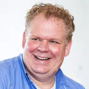 Lachende man met krullend blond haar in een blauw overhemd tegen een witte achtergrond.