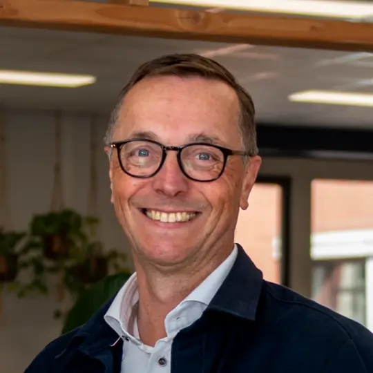 Lachende man met bril en donkergroen colbert in een lichte kamer met planten en ramen op de achtergrond.