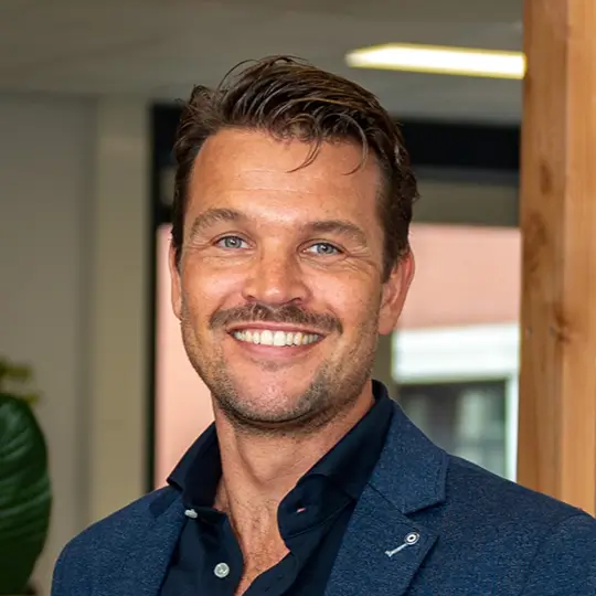 Een glimlachende man met blauw overhemd en donkerblauw colbert in een binnenruimte.