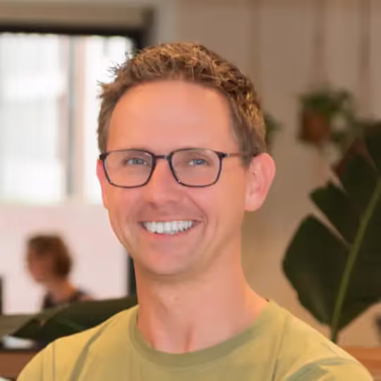 Man met bruine haar en bril glimlacht terwijl hij een groen shirt draagt, met een wazige achtergrond van planten en ramen.